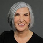 A woman with short gray hair and a black top smiles at the camera against a plain dark background.
