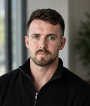 Man with short brown hair and beard wearing a black zip-up sweater sits indoors near large windows, looking at the camera.
