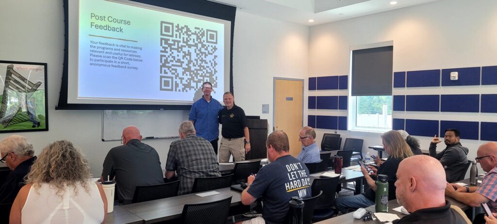 A group of adults sits in a classroom facing a screen displaying a large QR code and a post-course feedback message, while two men stand at the front.