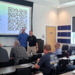 A group of adults sits in a classroom facing a screen displaying a large QR code and a post-course feedback message, while two men stand at the front.