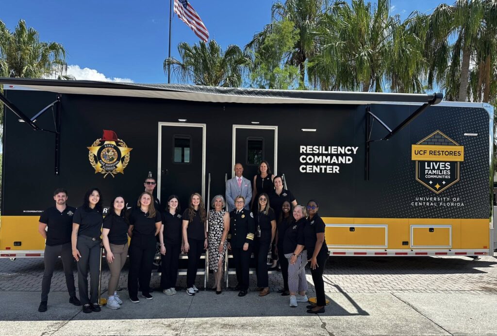 A group of people poses in front of a large black and yellow Resiliency Command Center vehicle with UCF Restores branding and palm trees in the background.