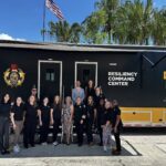 A group of people poses in front of a large black and yellow Resiliency Command Center vehicle with UCF Restores branding and palm trees in the background.