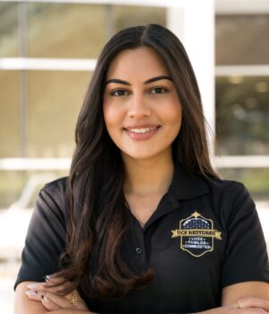 Woman with long dark hair stands outside a building, smiling with arms crossed, wearing a black polo shirt with a gold and blue embroidered badge.