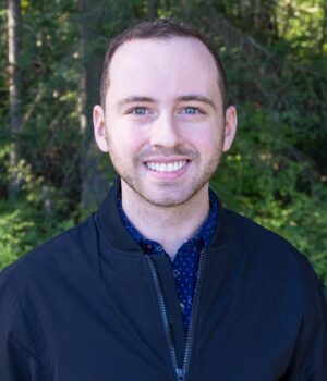 A person with short brown hair and a trimmed beard, wearing a black jacket, stands outdoors in front of green trees, smiling at the camera.